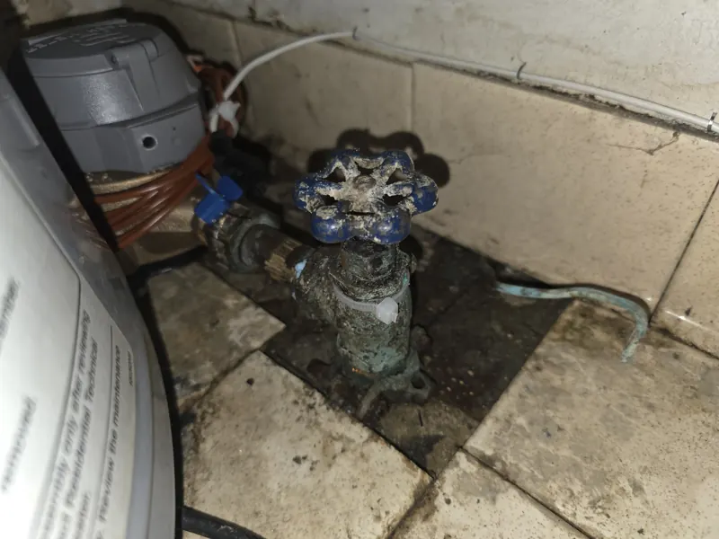 An old main water shut-off with a corroded meter yoke at a basement wall, water on the floor